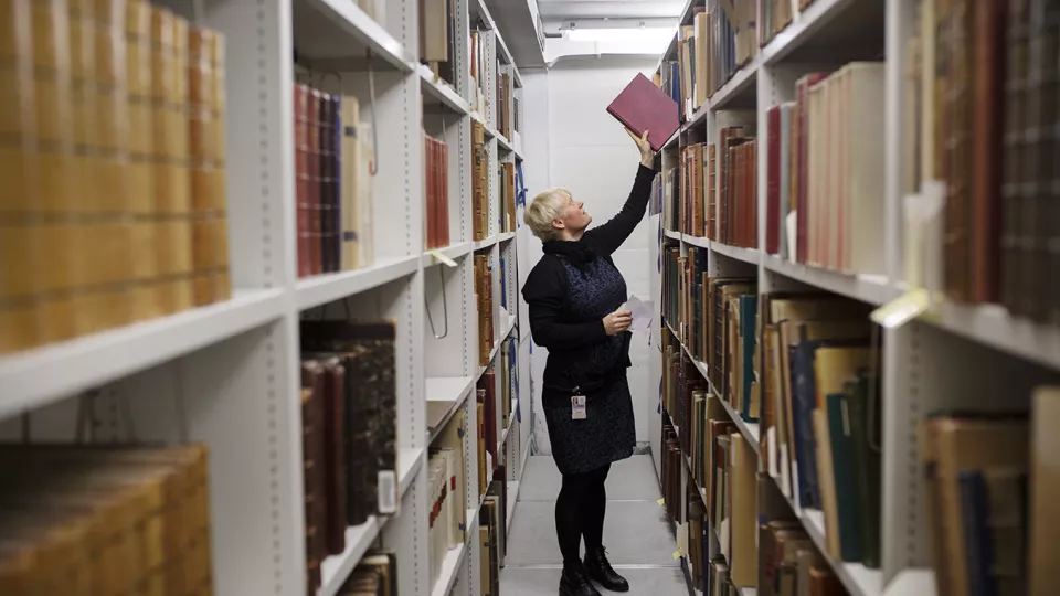 A librarian is standing between rows of books, picking down one.