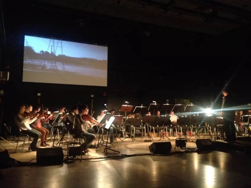 Orchestra infront of film on screen with boat in river