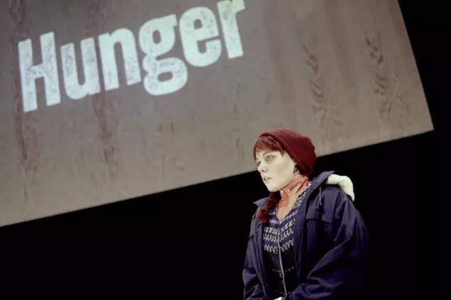 Kvinna i mössa och jacka framför en träskiva med texten Hunger