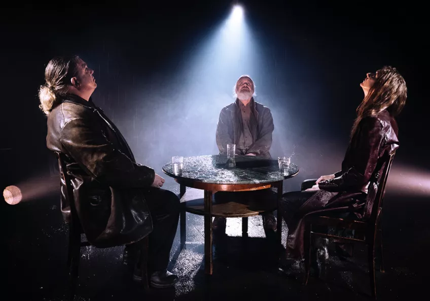 Tre personer sitter kring ett bord, i en teaterföreställning. 